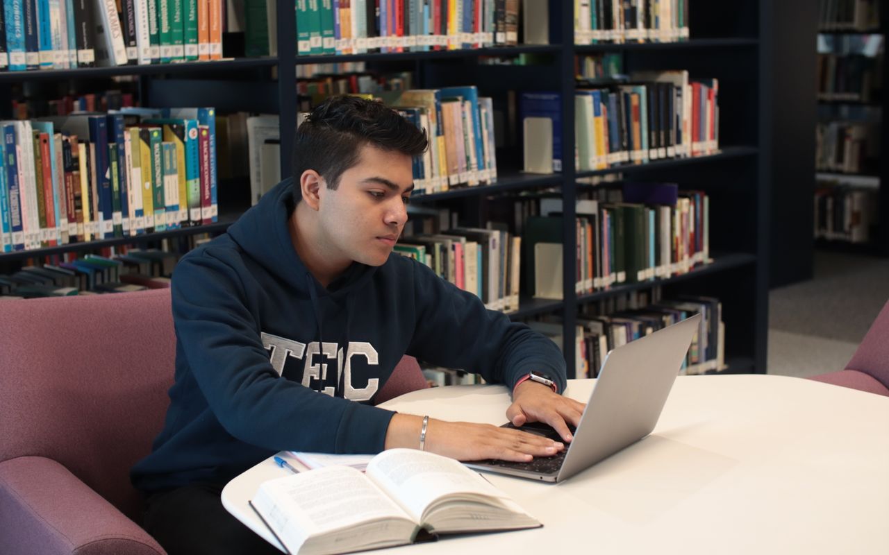 Estudante pesquisando em um computador entre livros de uma biblioteca